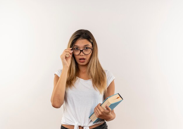 Chica estudiante bonita joven sorprendida con gafas y bolsa trasera sosteniendo el libro y sus gafas aisladas en la pared blanca