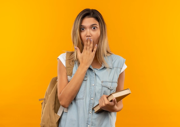 Chica estudiante bonita joven sorprendida que lleva el bolso trasero que sostiene el libro y que pone la mano en la boca aislada en la pared naranja