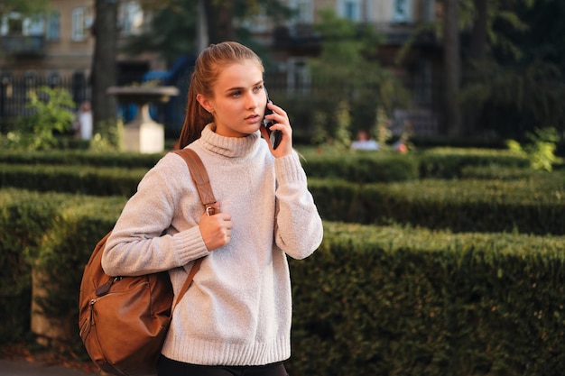 Chica estudiante casual atractiva en suéter acogedor con mochila hablando cuidadosamente por teléfono celular al aire libre