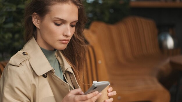 Chica estudiante concentrada que usa un teléfono inteligente para estudiar durante el descanso para tomar café en un café de la calle