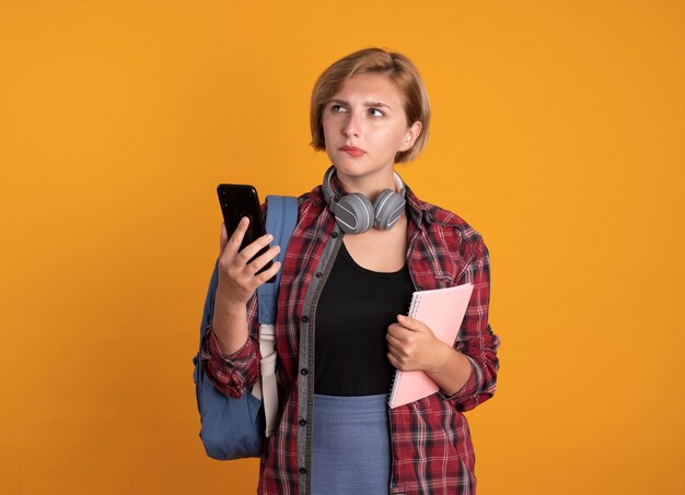 Chica estudiante eslava joven pensativa con auriculares con mochila tiene portátil y teléfono mirando al lado