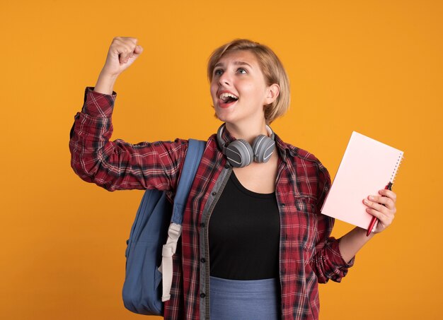 Chica estudiante eslava joven sorprendida con auriculares con mochila se encuentra con el puño levantado sostiene el cuaderno y la pluma mirando al lado