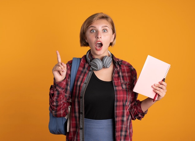 Chica estudiante eslava joven sorprendida con auriculares con mochila sostiene cuaderno y bolígrafo apunta hacia arriba