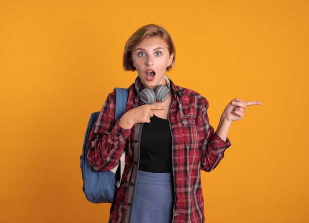 Chica estudiante eslava joven sorprendida con auriculares con puntos de mochila al lado con las dos manos