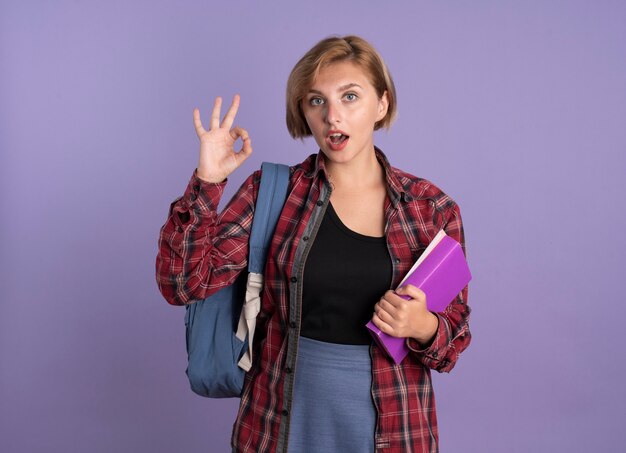 Chica estudiante eslava joven sorprendida con mochila sostiene gestos de libro y cuaderno bien signo de mano