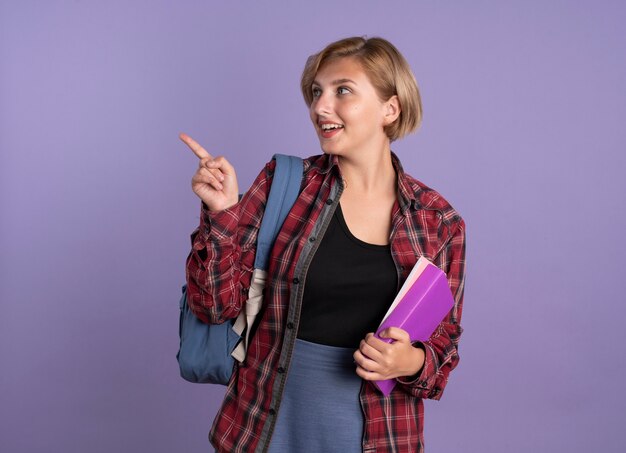 Chica estudiante eslava joven sorprendida con mochila sostiene el libro y el cuaderno mirando y apuntando al lado