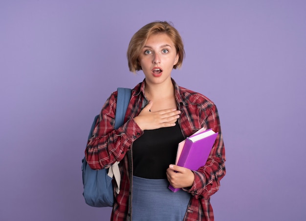 Chica estudiante eslava joven sorprendida con mochila sostiene el libro y el cuaderno pone la mano en el pecho