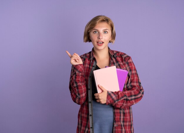 Chica estudiante eslava joven sorprendida sostiene puntos de libro y cuaderno al lado