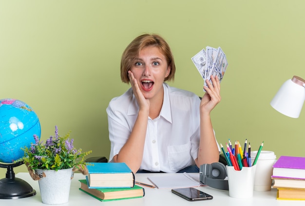 Chica estudiante rubia joven sorprendida sentada en el escritorio con herramientas escolares manteniendo la mano en la cara sosteniendo dinero