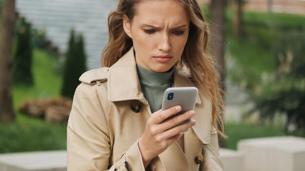 Chica estudiante rubia molesta que se siente triste enviando mensajes de texto con amigos en el chat en línea en el teléfono inteligente al aire libre Expresión de la cara