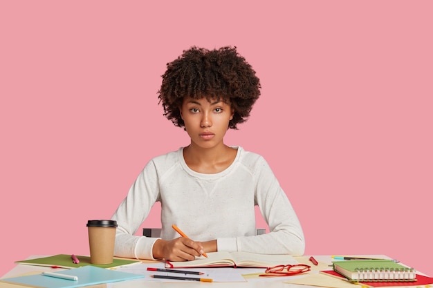 Chica estudiante seria concentrada posando en el escritorio contra la pared rosa