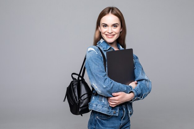 Chica estudiante sonriendo a la cámara, presionando la computadora portátil contra el pecho, con mochila, lista para ir a estudios, comenzar un nuevo proyecto y sugerir nuevas ideas aisladas en la pared gris