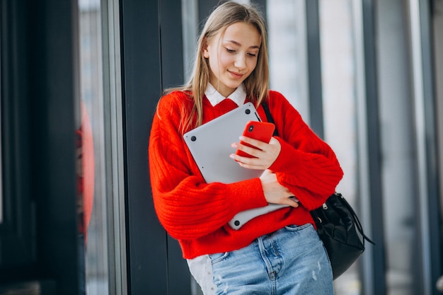 Chica estudiante sosteniendo la computadora y hablando por teléfono junto a la ventana