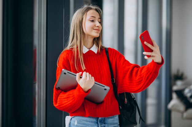 Chica estudiante sosteniendo la computadora y hablando por teléfono junto a la ventana