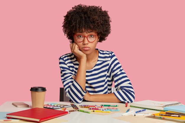 Chica estudiante triste posando en el escritorio contra la pared rosa