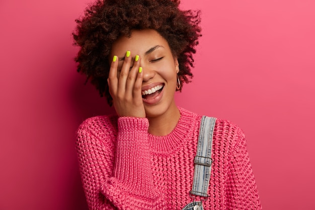 Chica étnica despreocupada no puede dejar de reír, mantiene la mano en la cara, tiene cara alegre, sonríe positivamente, tiene buen sentido del humor, expresa felicidad, usa jersey de punto, posa sobre una pared rosa