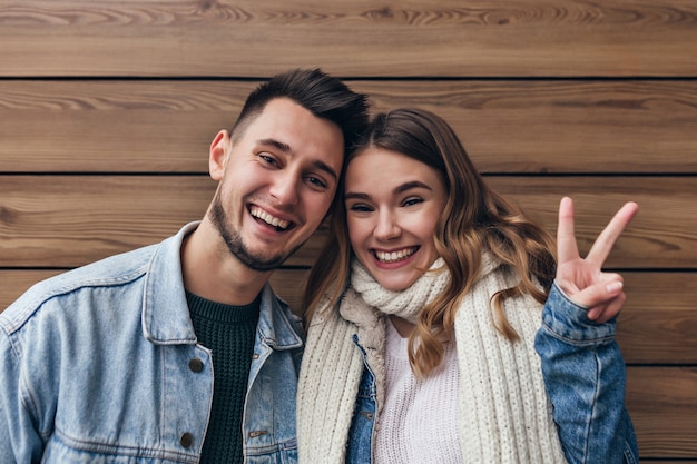 Chica europea alegre en bufanda tejida posando con el signo de la paz en la pared de madera. Filmación en interiores de la risa pareja casada divirtiéndose juntos.