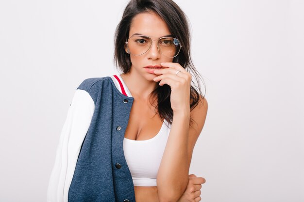 Chica europea bronceada desagradable en vasos tocando la mejilla con los dedos. Retrato de primer plano de mujer morena seria lleva posando anillo de plata.