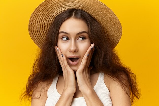 Chica europea delgada de moda positiva con moño suelto pelo ondulado con sombrero de paja mirando a otro lado