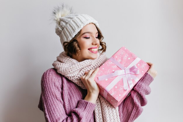 Chica europea de pelo corto con sombrero posando emocionalmente, mirando el presente de año nuevo. Foto interior de señorita sorprendida en lindo pañuelo con regalo rosa.