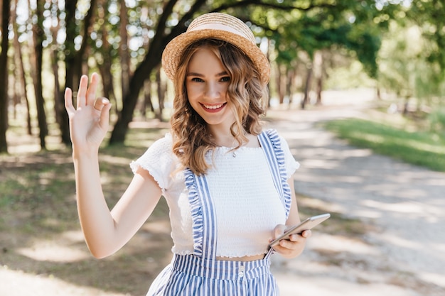 Chica europea con sombrero posando en el parque con expresión de cara feliz y agitando la mano. Modelo de mujer alegre en traje de verano vintage divirtiéndose.