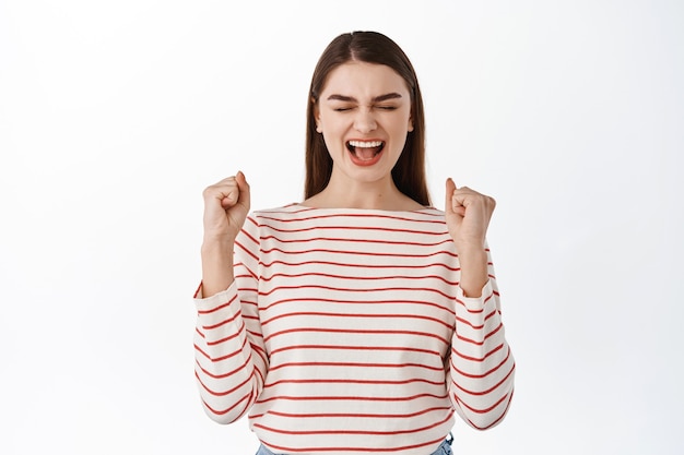 Chica exitosa emocionada celebrando el logro, ganando y gritando que sí, haciendo puñetazos, ganando su objetivo, triunfando de la victoria, de pie sobre una pared blanca