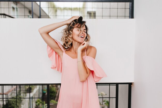 Chica extasiada en vestido rosa de verano posando con los ojos cerrados. Retrato de elegante modelo de mujer bronceada riendo en buen día.