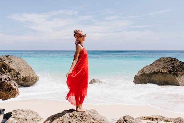 Chica extática con bonita sonrisa de pie sobre piedra grande con océano. Foto de cuerpo entero al aire libre de alegre turista relajándose en la playa salvaje.