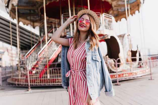 Chica extática viste chaqueta vaquera posando junto al carrusel con una sonrisa sincera. Foto al aire libre de linda mujer rubia en vestido de rayas pasando el día en el parque de atracciones.