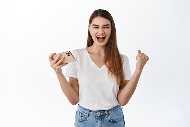 Chica extremadamente feliz grita sí y sostiene que el teléfono inteligente gana en el juego móvil logra el éxito de la red de objetivos celebrando la victoria de pie sobre fondo blanco