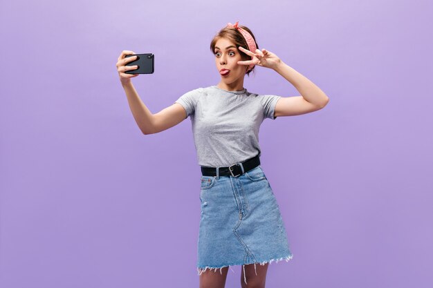 Chica en falda de mezclilla demuestra lengua, muestra el signo de la paz y se toma selfie. Mujer divertida de buen humor en ropa elegante posando.