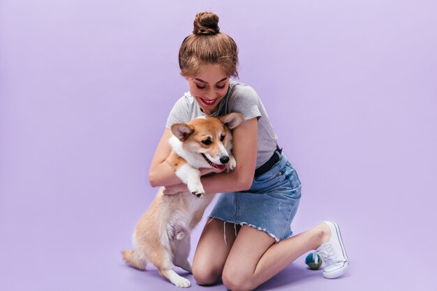 Chica en falda de mezclilla juega con corgi sobre fondo morado. Linda mujer joven en traje de moda tiene corgi sobre fondo aislado.