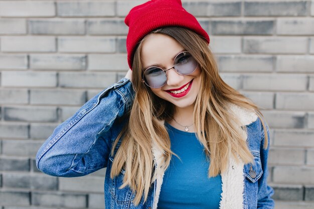 Chica fascinante con expresión facial complacida posando en la calle por la mañana. Foto exterior de guapa dama europea viste sombrero rojo sonriendo