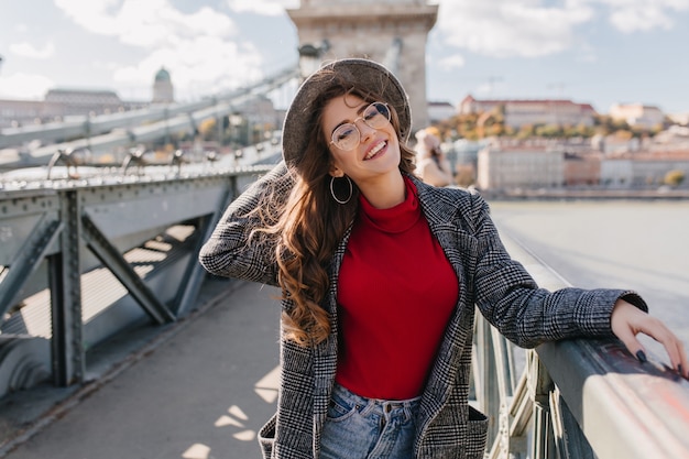 Chica fascinante en suéter rojo tejido sonriendo juguetonamente en el puente en un día soleado