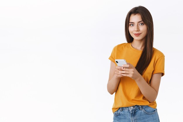 Chica feliz, atrevida y confiada en camiseta amarilla, sosteniendo un teléfono inteligente con una cámara asertiva sonriente, mensajería, usando una aplicación móvil, comprando en línea o navegando por Internet, de pie con fondo blanco.