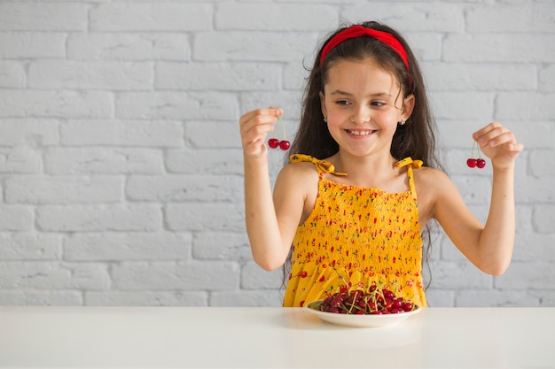 Chica feliz celebración de cerezas rojas en la mesa contra la pared de ladrillo blanco