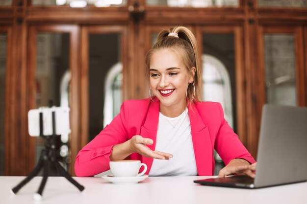 Foto gratuita chica feliz con chaqueta rosa con taza de café y laptop grabando alegremente un nuevo video para su vlog