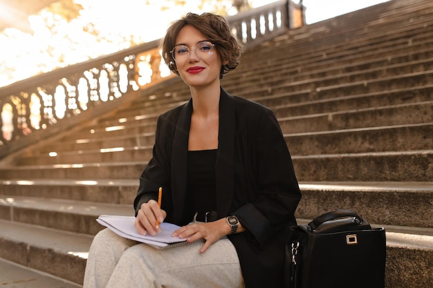 Chica feliz con chaqueta y top negro sonriendo afuera Mujer de cabello ondulado con labios rojos en anteojos se sienta en las escaleras al aire libre