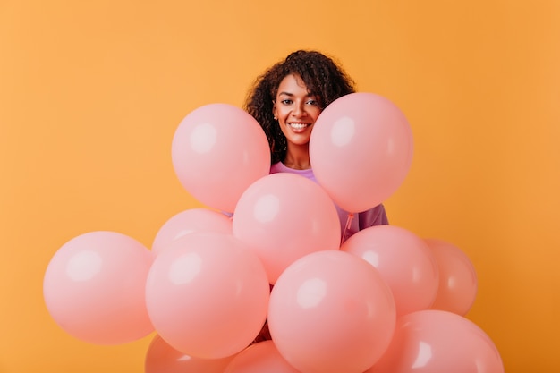 Chica feliz cumpleaños posando con sonrisa alegre. Retrato interior de una mujer bastante africana con globos de fiesta aislados en naranja.