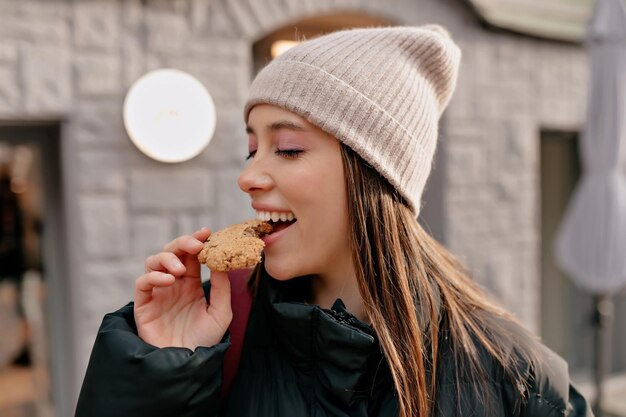Chica feliz y emocionada con cabello oscuro con gorra beige y chaqueta oscura está mordiendo galletas en el café al aire libre bajo el sol
