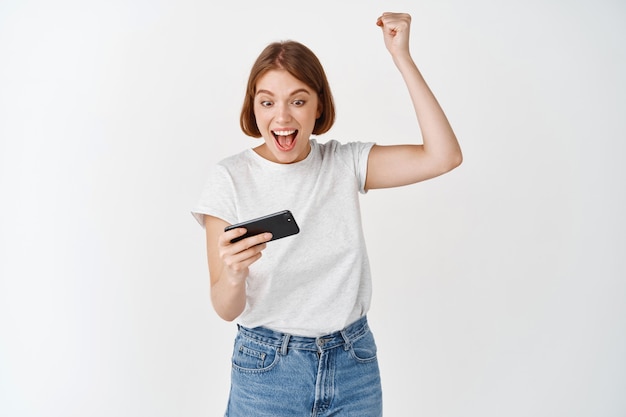 Chica feliz ganando videojuegos, levantando la mano y mirando el teléfono inteligente emocionado, celebrando la victoria en la aplicación móvil, pared blanca