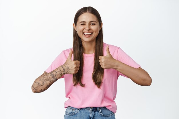 Chica feliz guiñando un ojo y mostrando los pulgares hacia arriba, sonriendo con dientes blancos, con camiseta rosa, fondo blanco.