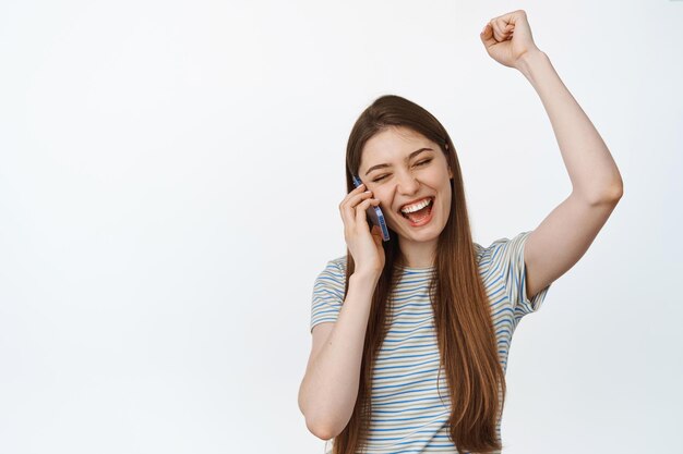 Chica feliz hablando por teléfono móvil y celebrando. Una mujer emocionada recibe buenas noticias de guardia, de pie contra un fondo blanco.