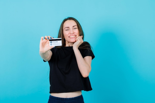 La chica feliz muestra la tarjeta de crédito a la cámara y sostiene la otra mano en la mejilla con fondo azul.