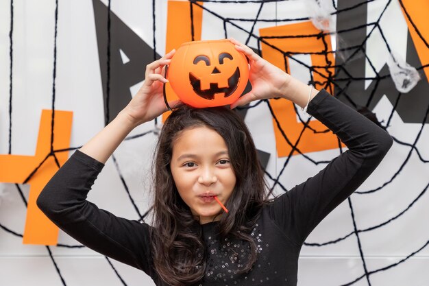 Chica feliz de pie frente a la decoración de la fiesta de Halloween y sosteniendo calabaza Jacko'lantern sobre la cabeza