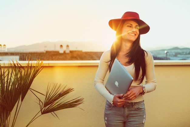 Foto gratuita chica feliz posando con un sombrero y un portátil al atardecer