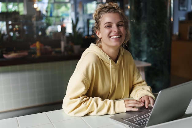 Chica feliz sentada en un café y trabajando con una computadora portátil Mujer alegre sonriente usando una computadora portátil en una cafetería Freelancer en espacio de coworking Estudiante estudiando en línea en un café