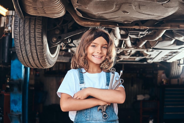 Una chica feliz y sonriente está parada debajo del auto en el taller de autos con una llave en las manos.
