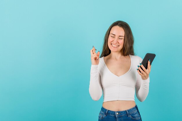 La chica feliz sostiene el teléfono inteligente y levanta la otra mano cruzando los dedos en el fondo azul