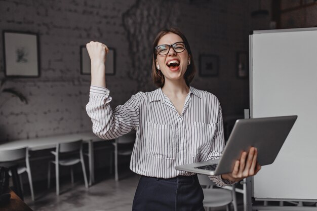 Chica felizmente grita y hace un gesto con la mano ganadora, sosteniendo la computadora portátil y posando en la oficina contra el fondo del tablero.